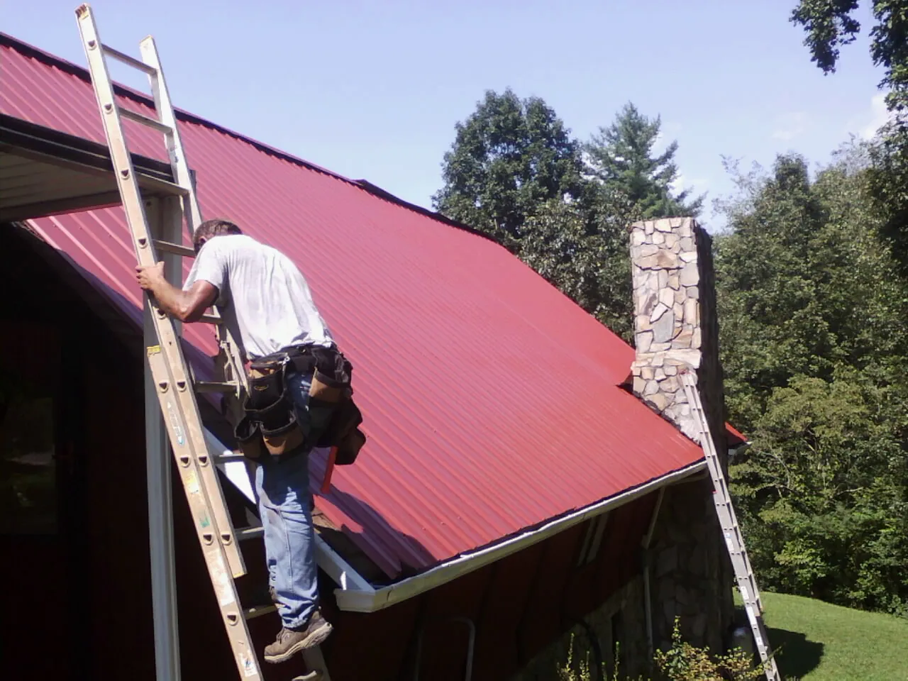 Roofer on a ladder beside a red metal roof and stone chimney — Soffit & Fascia Repair by Roofing Brothers in Markleton